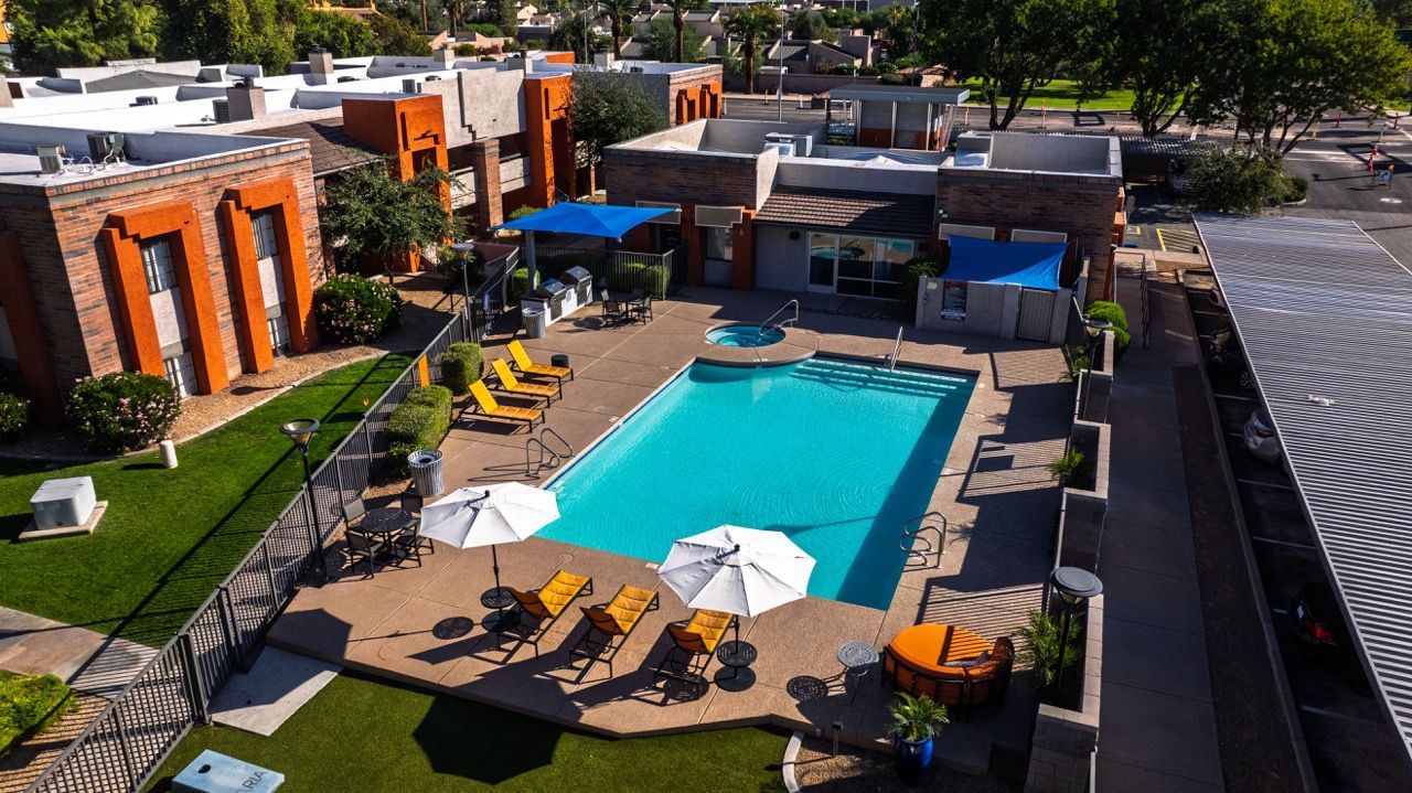 Aerial view of apartment complex pool with lounge chairs and shade umbrellas; buildings in the background.
