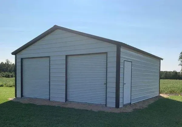 A white garage with two garage doors is sitting in the middle of a grassy field.