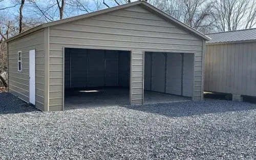 A garage with two doors is sitting on top of a gravel driveway next to a house.