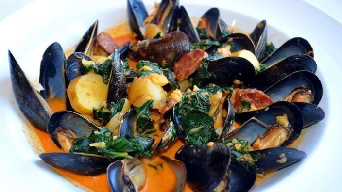 Mussels in a bright orange broth with potatoes, spinach, and chorizo in a white bowl.