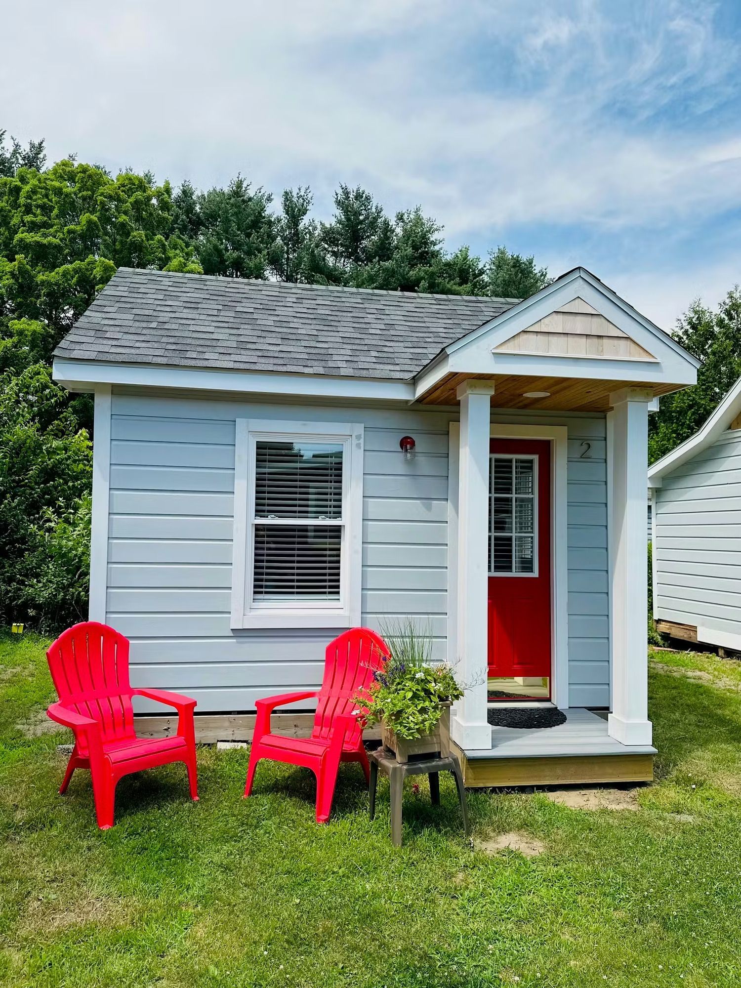 Small light blue cabin with red door, chairs, and potted plant on a grassy lawn.