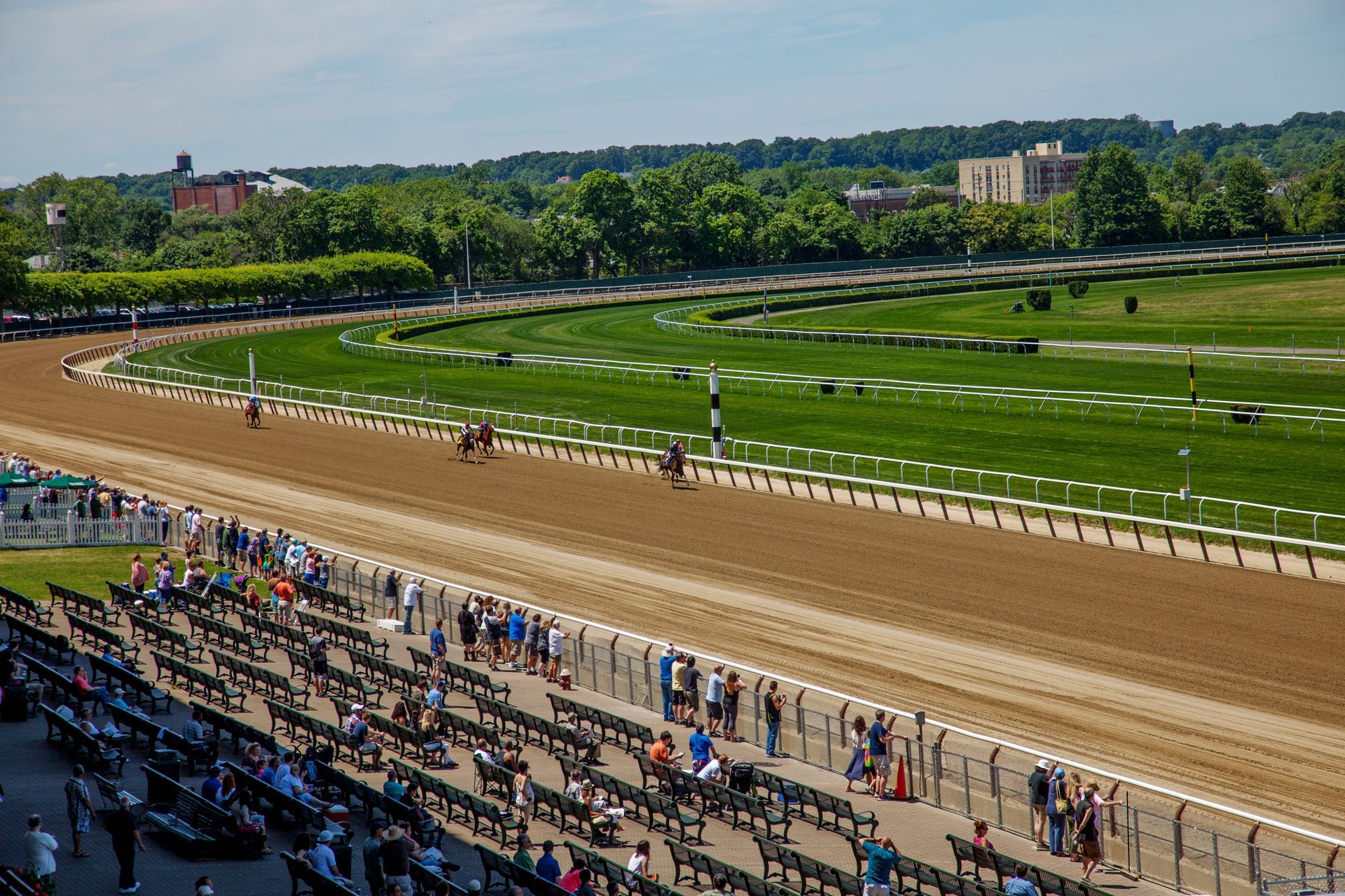 Horse race at a racetrack. Brown track, green field, spectators in the stands, trees in the background.
