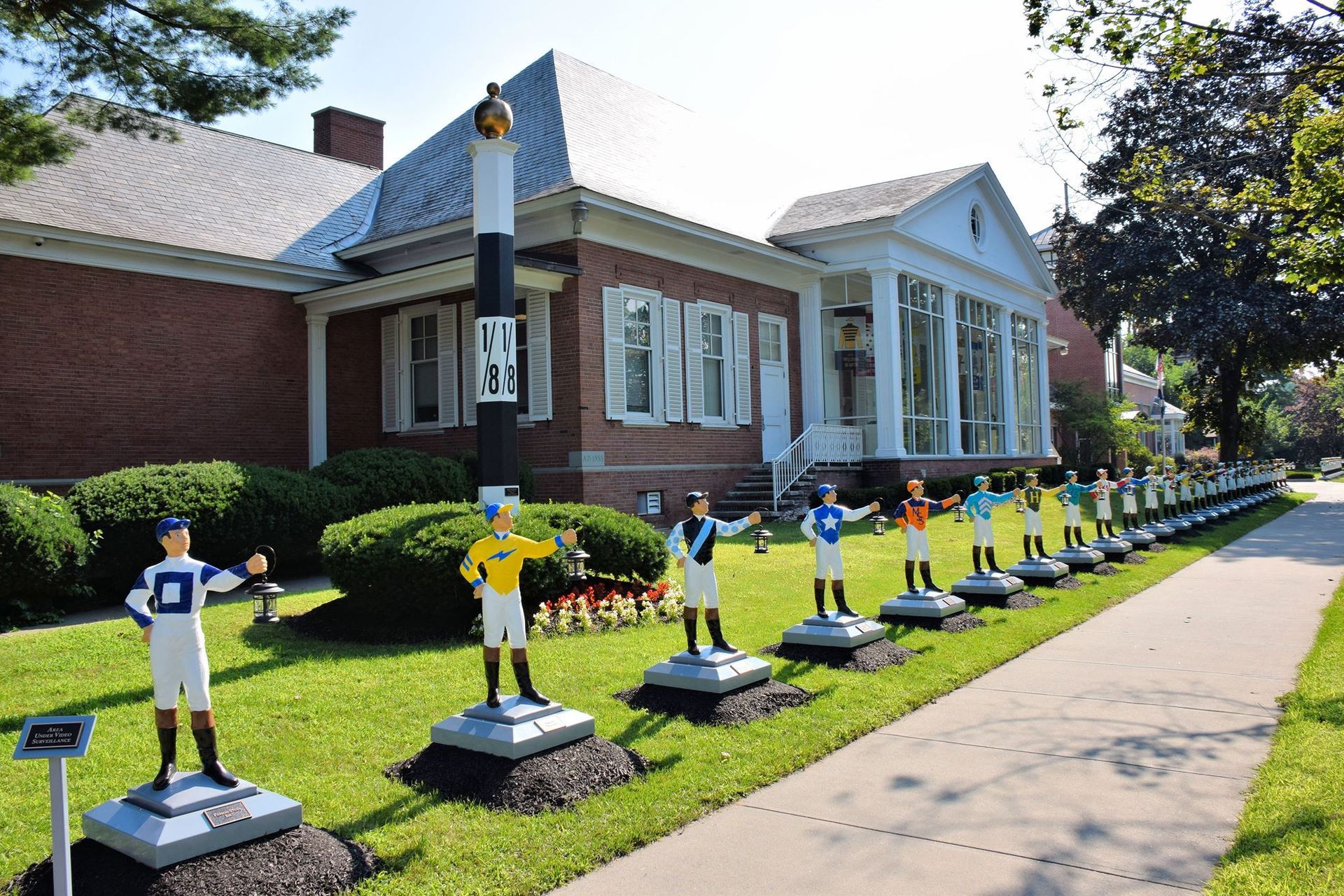 Lined statues of jockeys in various colors stand on a lawn; brick house with columns in background.