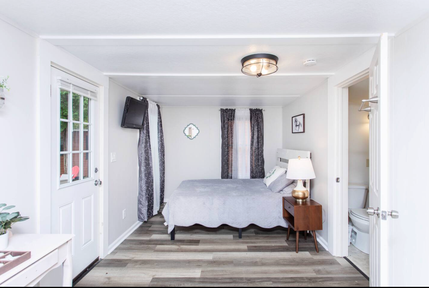 Small bedroom with bed, nightstand, door, window, and TV. Grey and white color scheme.