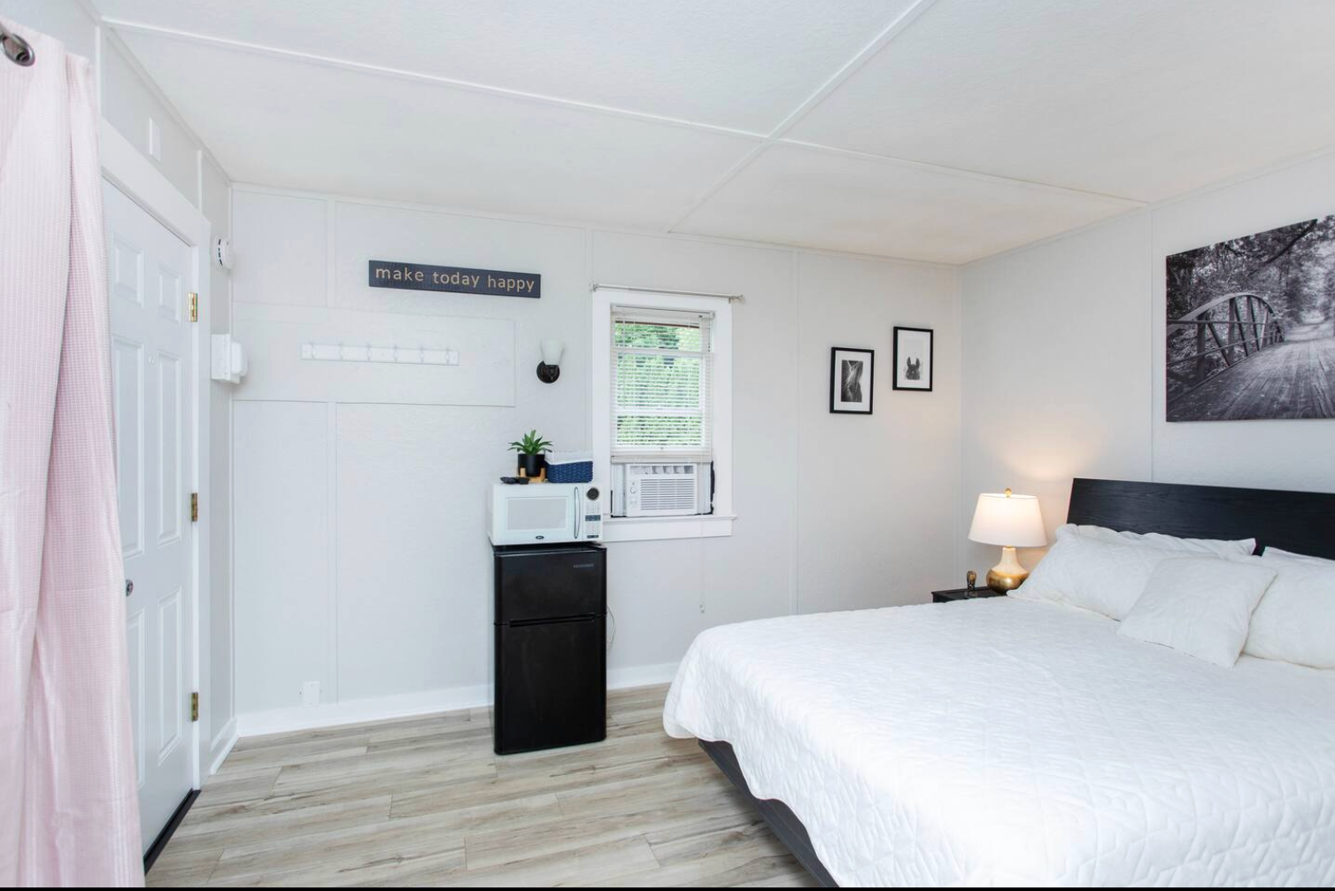 Small bedroom with bed, mini-fridge, microwave, window, art, and a closed door with pink curtain.