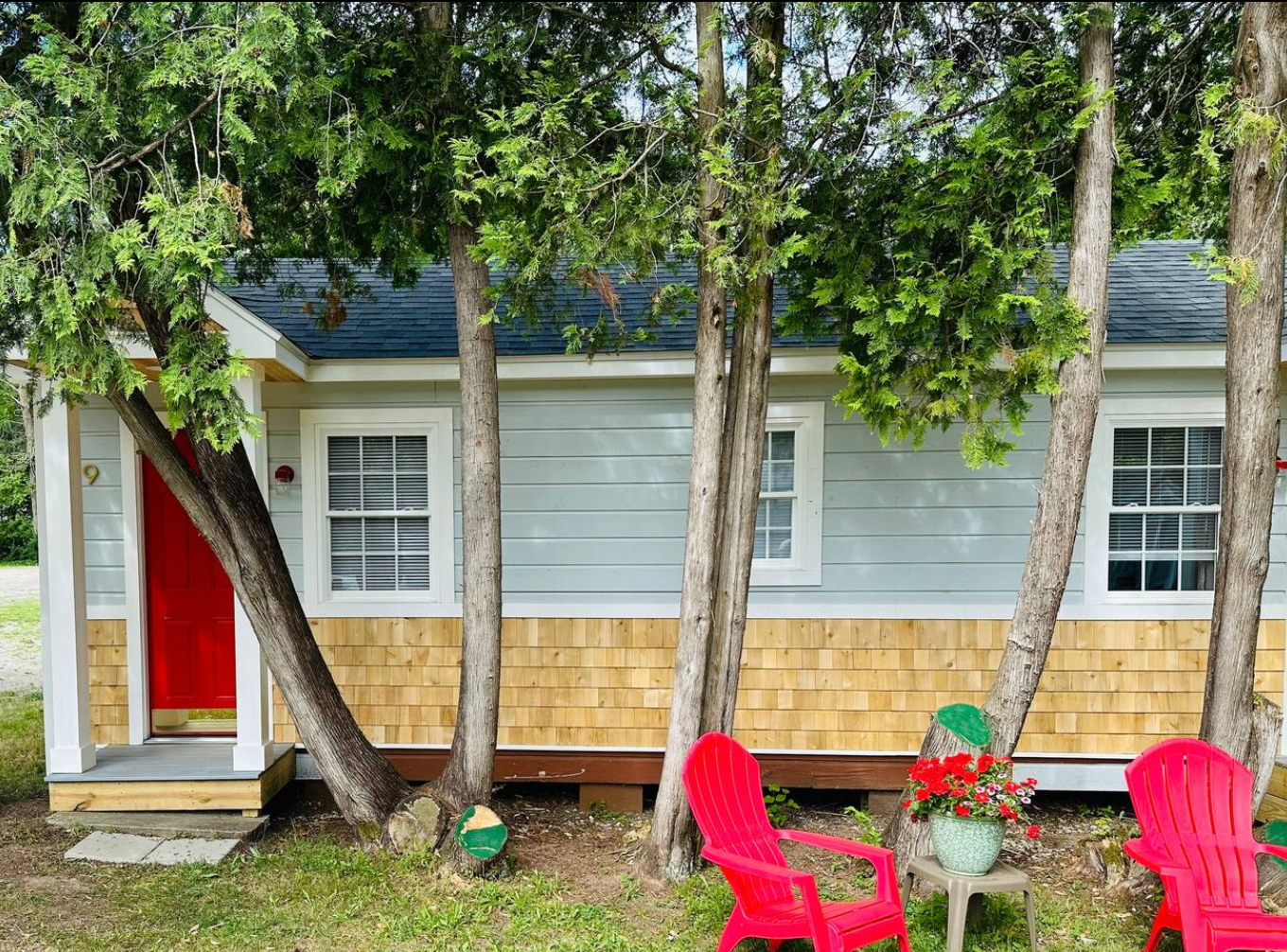 Small light blue cottage with cedar shake siding, red door, and red chairs.