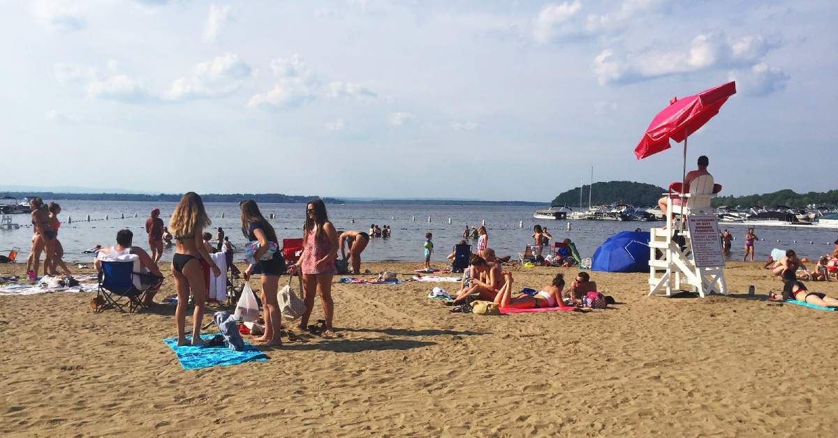 Beach scene with people, lifeguard tower, red umbrella, and boats on the water.