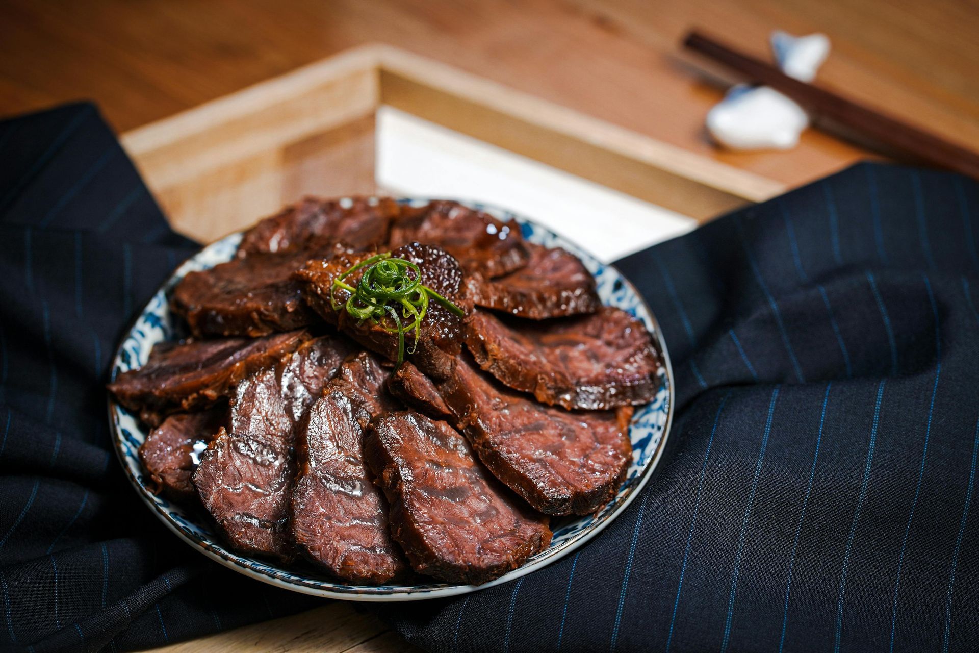 Sliced, glazed meat on a patterned plate, garnished with green herbs, displayed on a wooden tray with chopsticks.