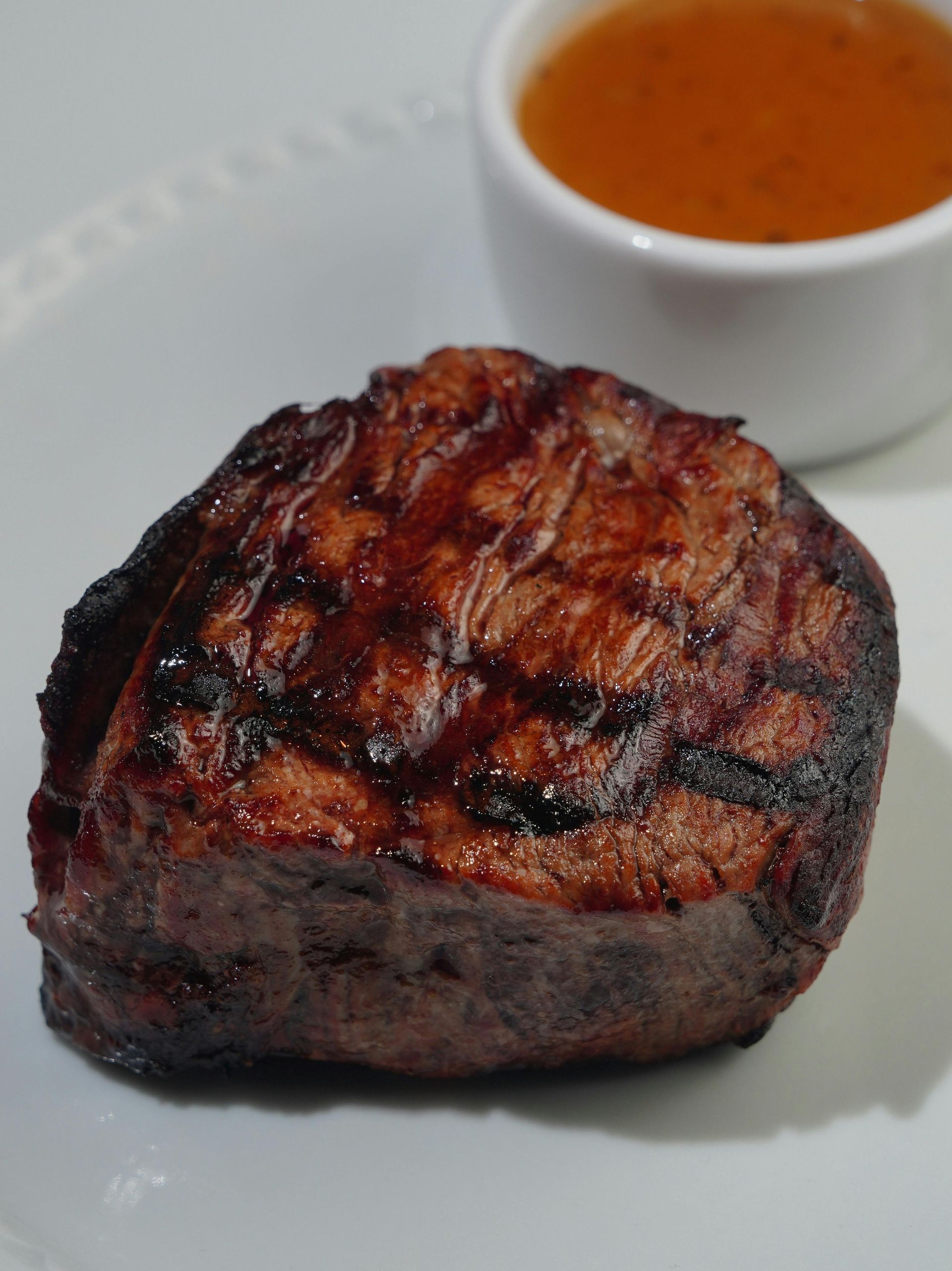 Grilled steak with sauce in a small white cup on a white plate.