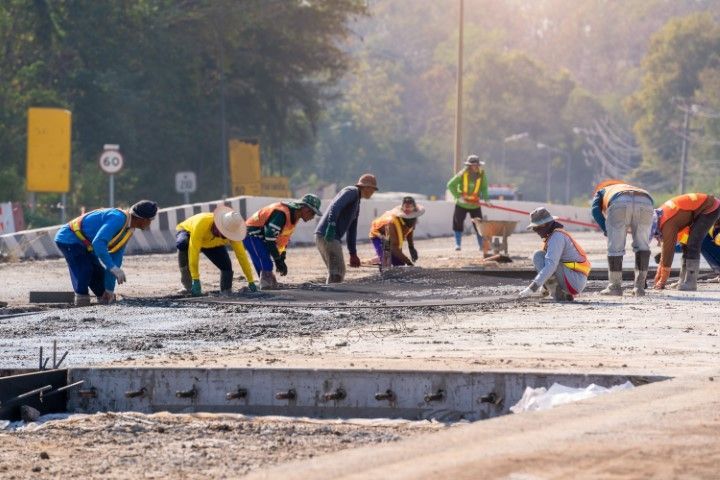 Construction workers paving a road, bending over wet concrete with tools at a roadside site.