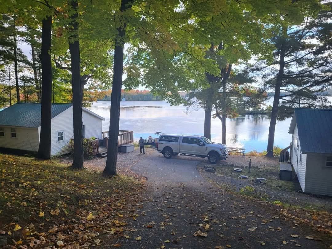 Wooded lakeside driveway with parked SUV, small cabins, and misty water in the background