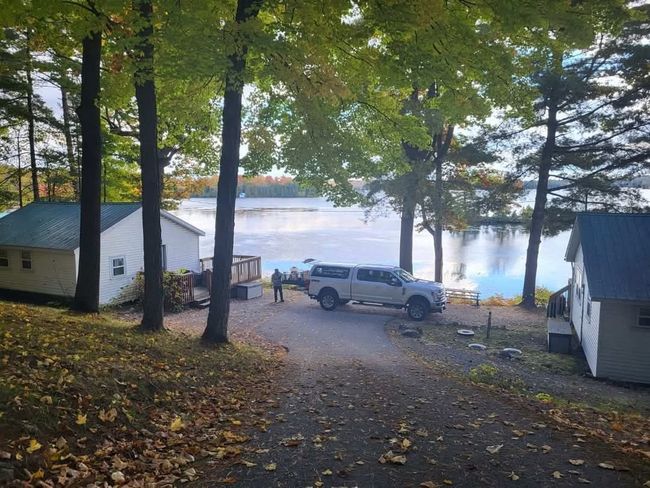Wooded lakeside driveway with parked SUV, small cabins, and misty water in the background
