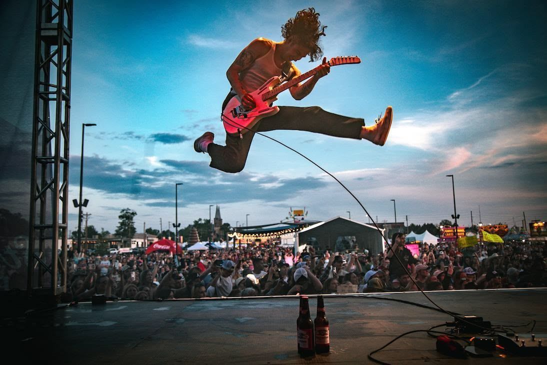 A performer jumps mid-air while playing a bright pink electric guitar on an outdoor concert stage.