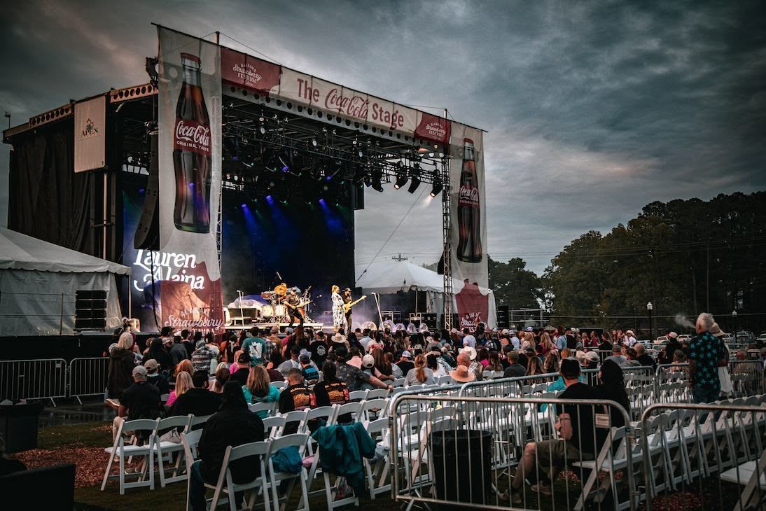 A live outdoor music concert at dusk with a band performing on stage before an audience seated in folding chairs.