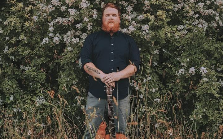 A man with a beard is standing in a field holding a guitar.