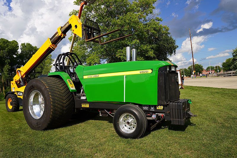 A modified green and yellow John Deere pulling tractor with a bright yellow forklift attachment on a grassy field.