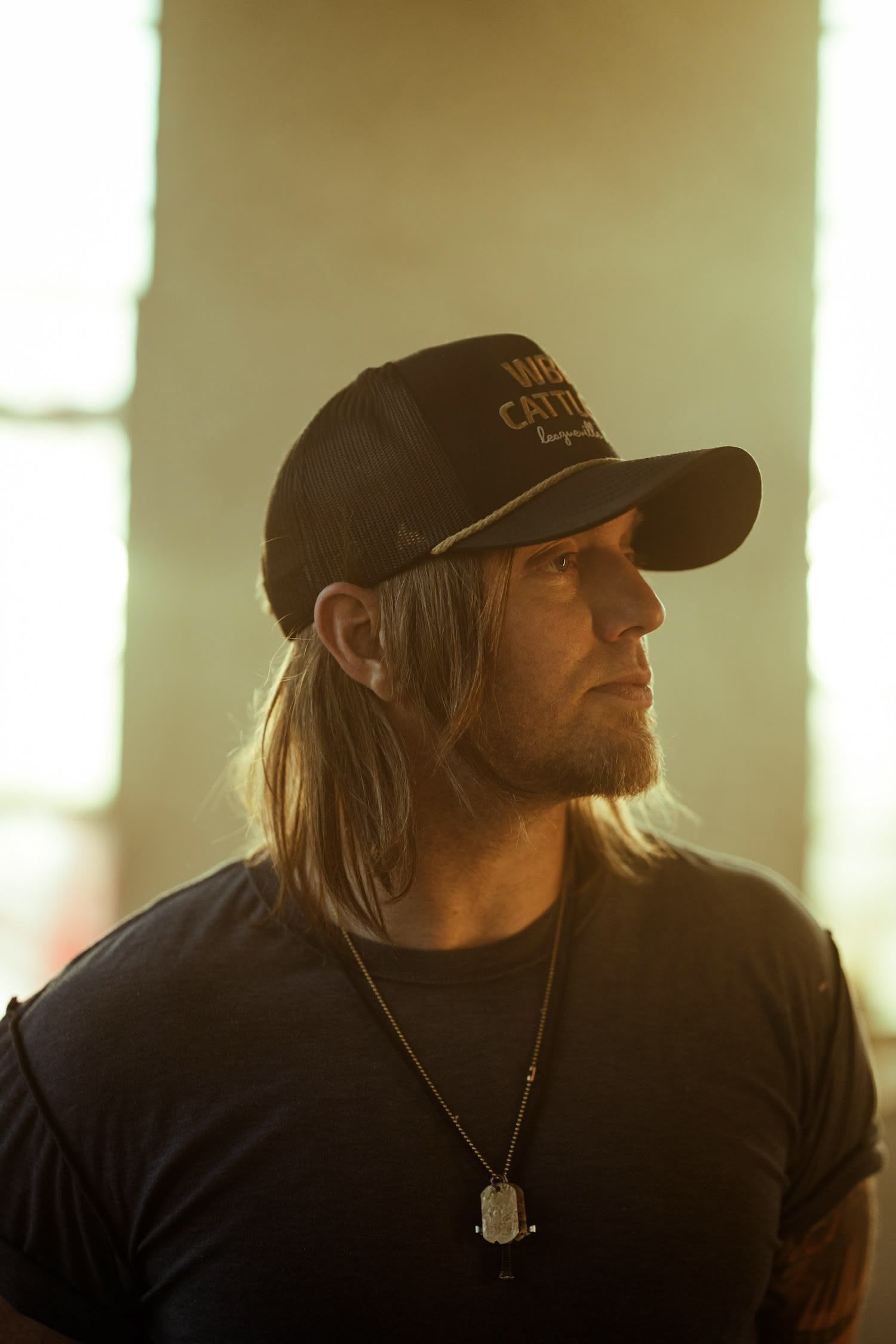 A man wearing a hat and a necklace is standing in front of a window.