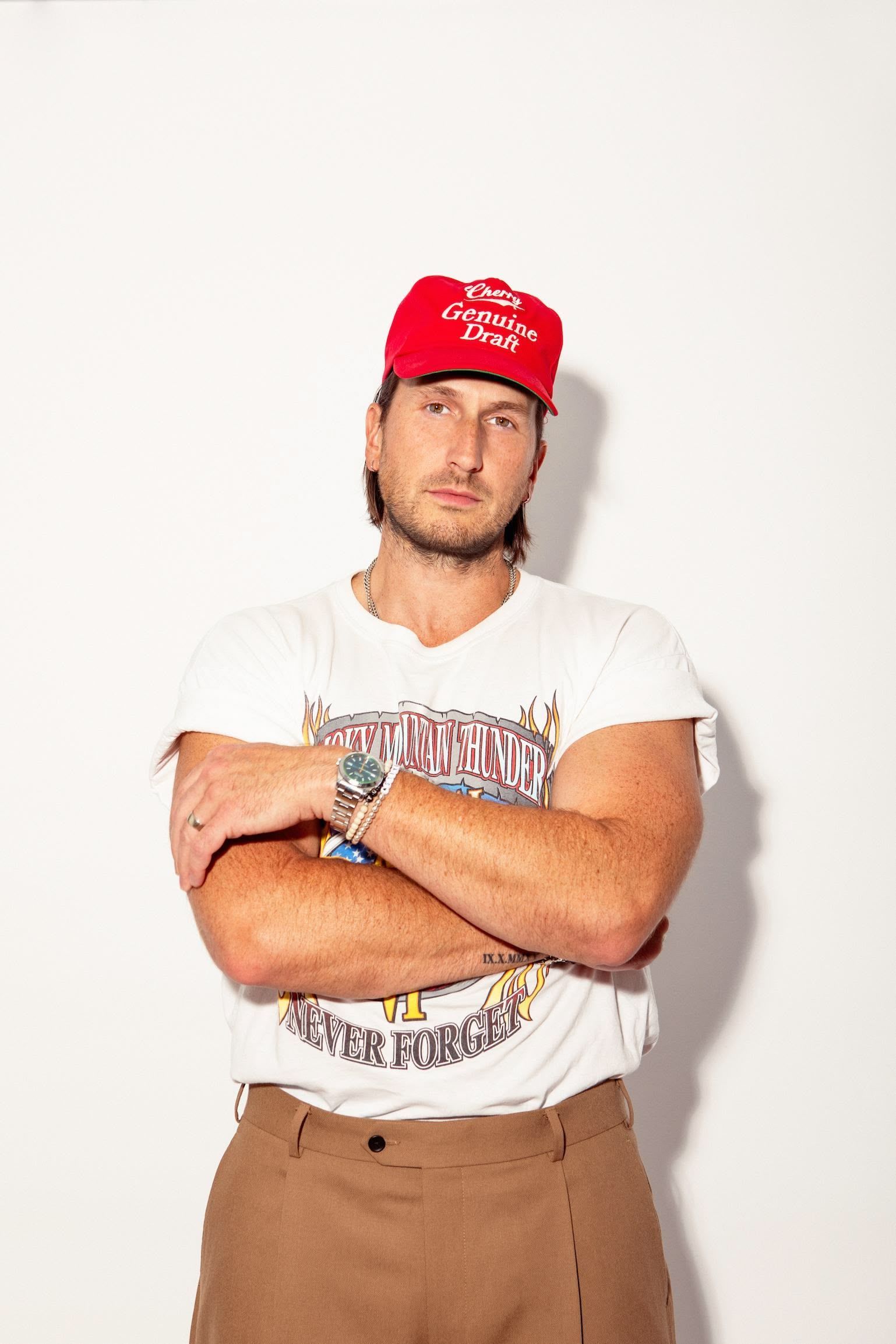 A man wearing a red hat and a white t-shirt is standing with his arms crossed.
