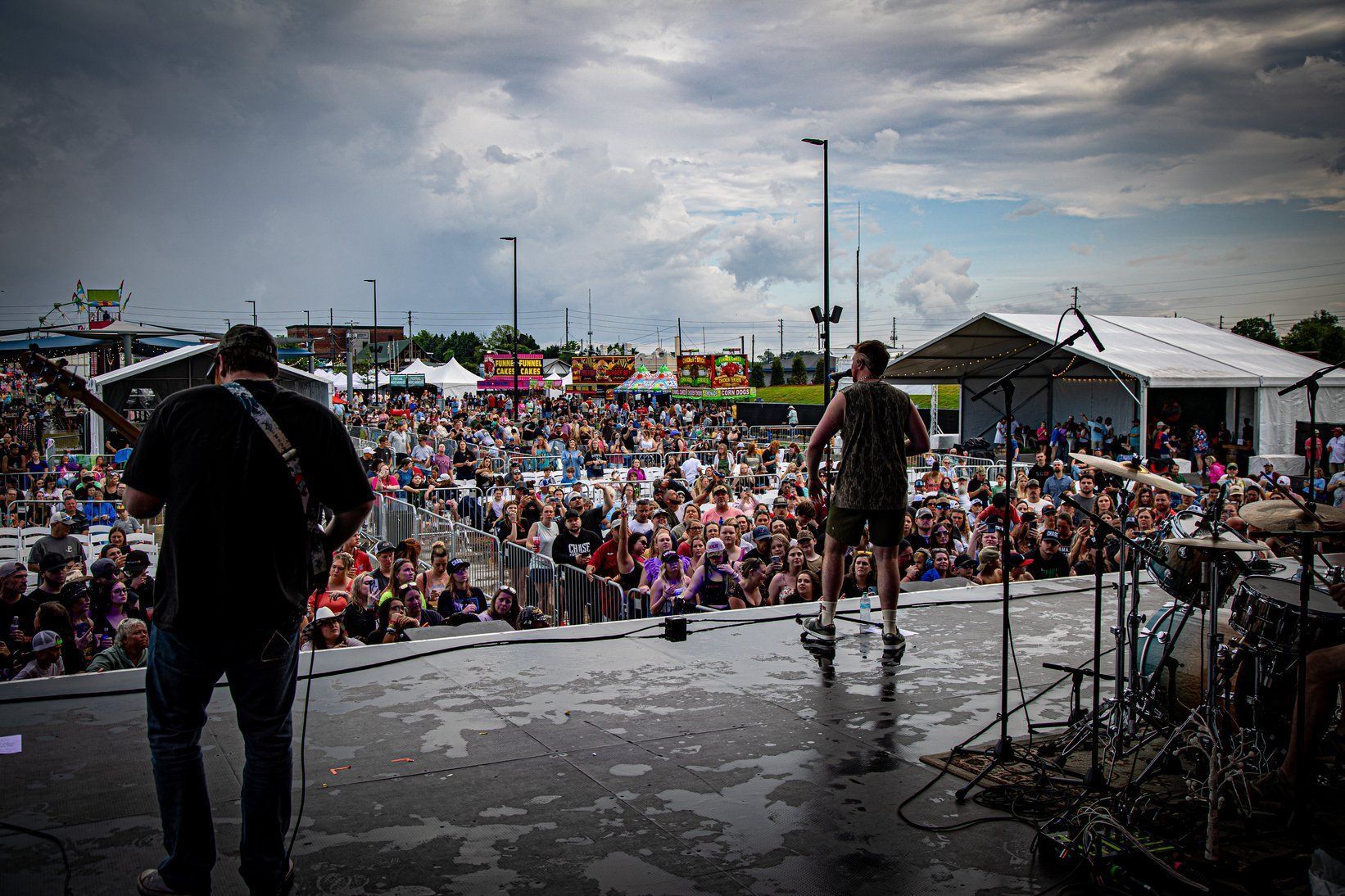 Two performers stand on a stage before a large, distant crowd at an outdoor summer festival under a cloudy sky.
