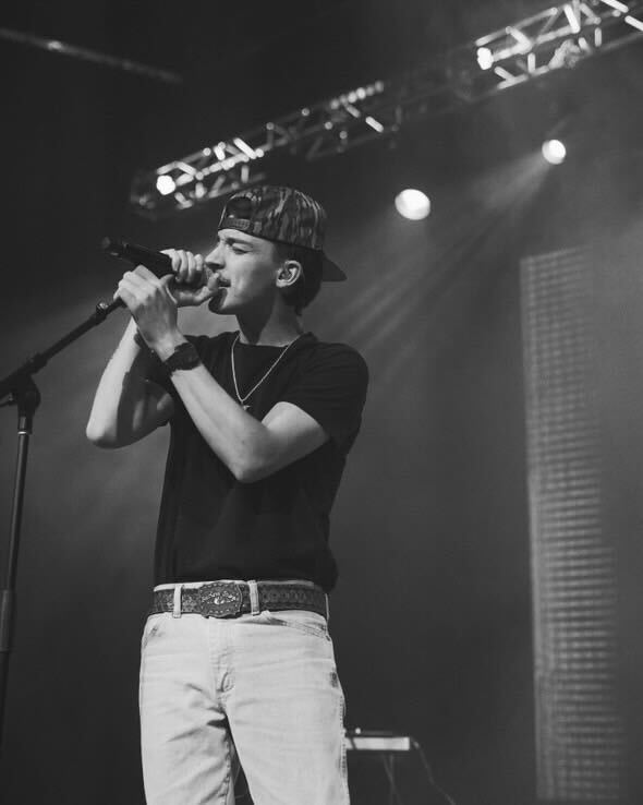 A man wearing a hat and a necklace is standing in front of a window.