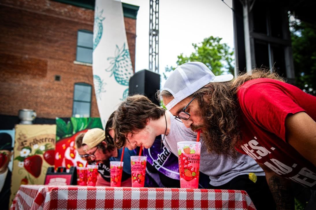 People leaning over a checkered table to drink from cups with straws during an outdoor competition.