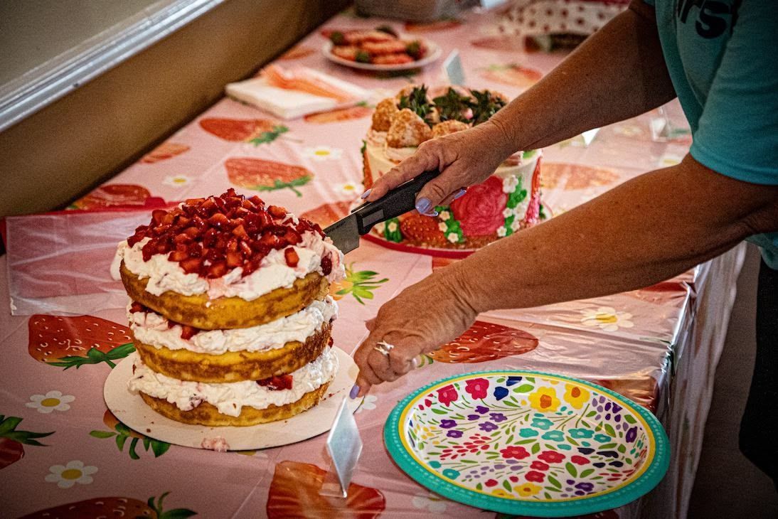 A person slices a three-layer strawberry cake on a floral tablecloth next to an empty plate.