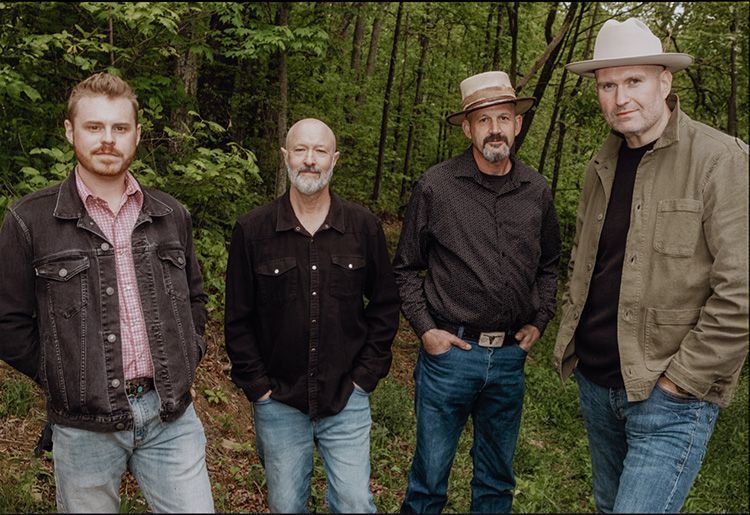 Four men pose outdoors in a wooded area. They wear casual attire, including denim and hats.