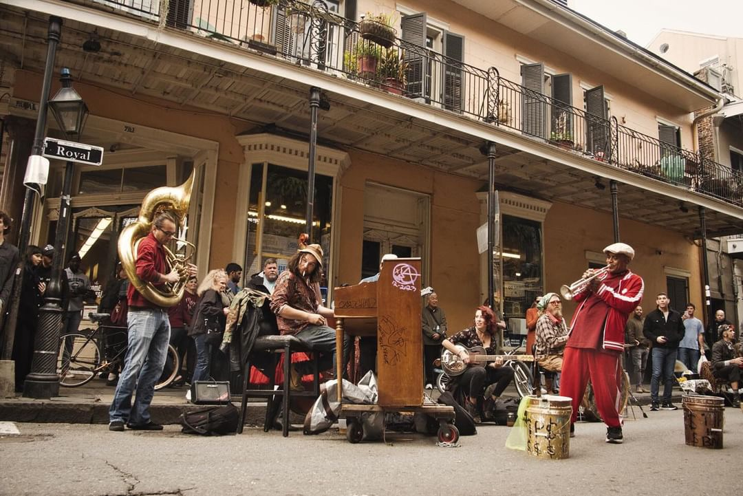 Street musicians playing music in New Orleans: tuba, keyboard, drums, flute. Building with balcony in background.