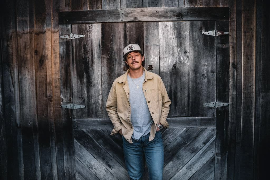 A man wearing glasses and a hat is kneeling down with his hands on his hips.