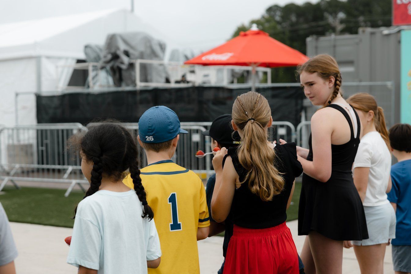 A group of individuals in casual clothing stand in line outdoors near a metal fence, a tent, and a red umbrella.