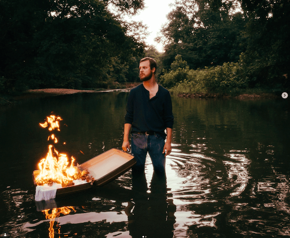A man wearing glasses and a hat is kneeling down with his hands on his hips.