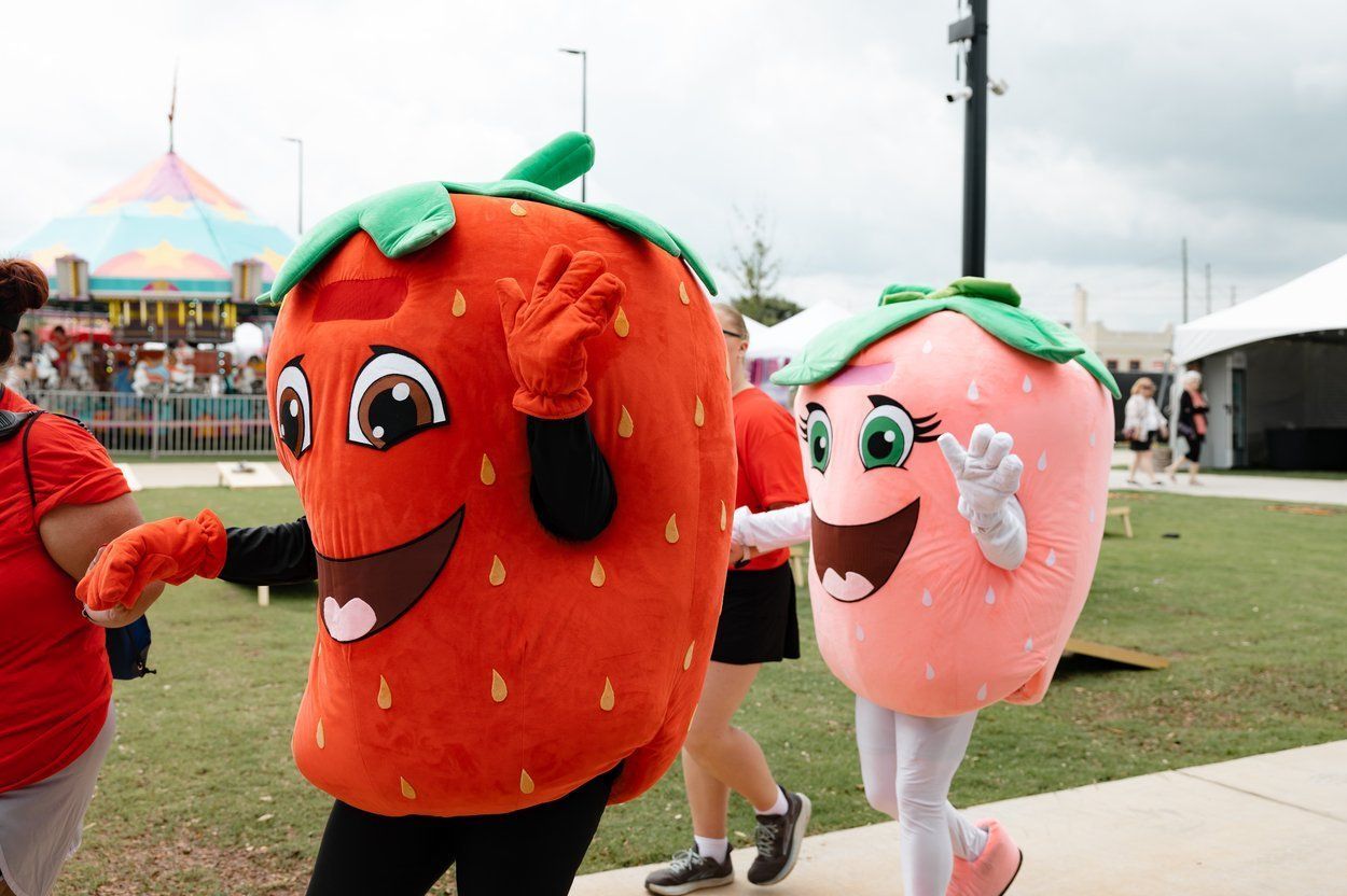 Two strawberry mascots, one bright red and one light pink, walk outdoors at a fair with a tent in the background.