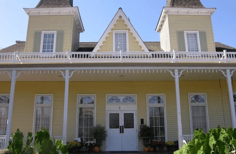 A light yellow, two-story building with a wide front porch, two tower-like roof peaks, and a white double-door entrance.