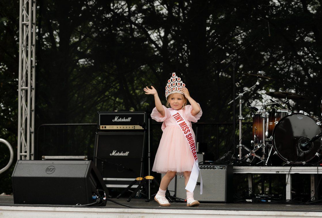 A child wearing a tiara and a pink dress with a sash waves from a stage in front of musical equipment.