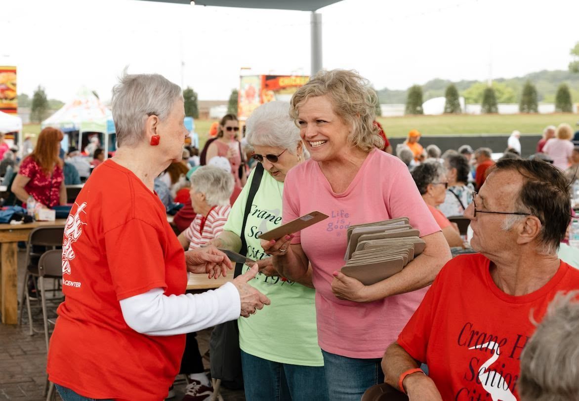 People in casual clothing interact at an outdoor gathering, with some holding documents under a tent canopy.