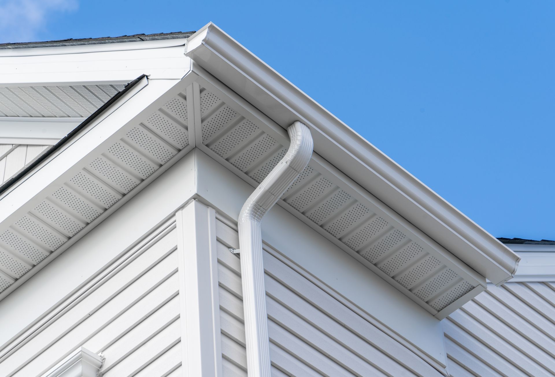 White house corner with vinyl siding, roof eaves, and a downspout against a blue sky