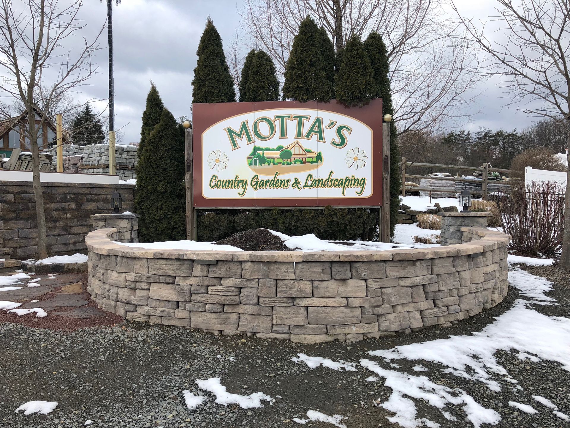 A sign for motta 's country gardens and landscaping is sitting on top of a stone wall.