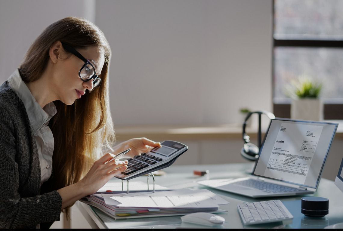 A woman is sitting at a desk using a calculator and a laptop.