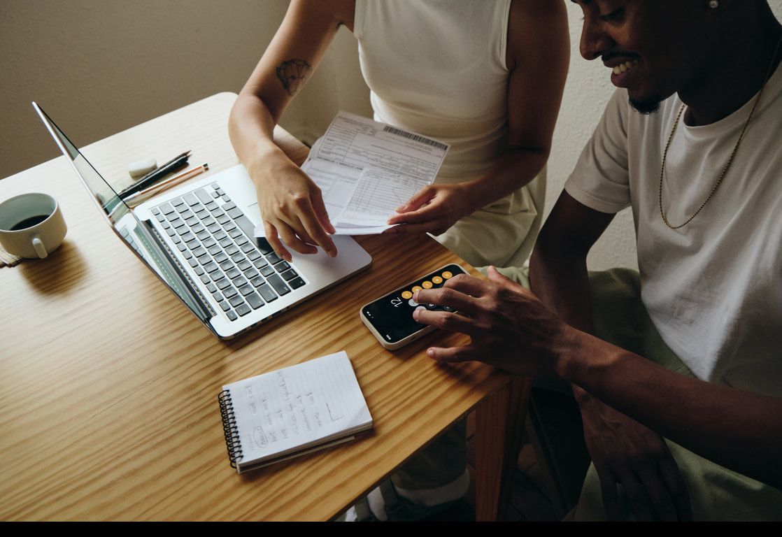A man and a woman are sitting at a table with a laptop and a cell phone.