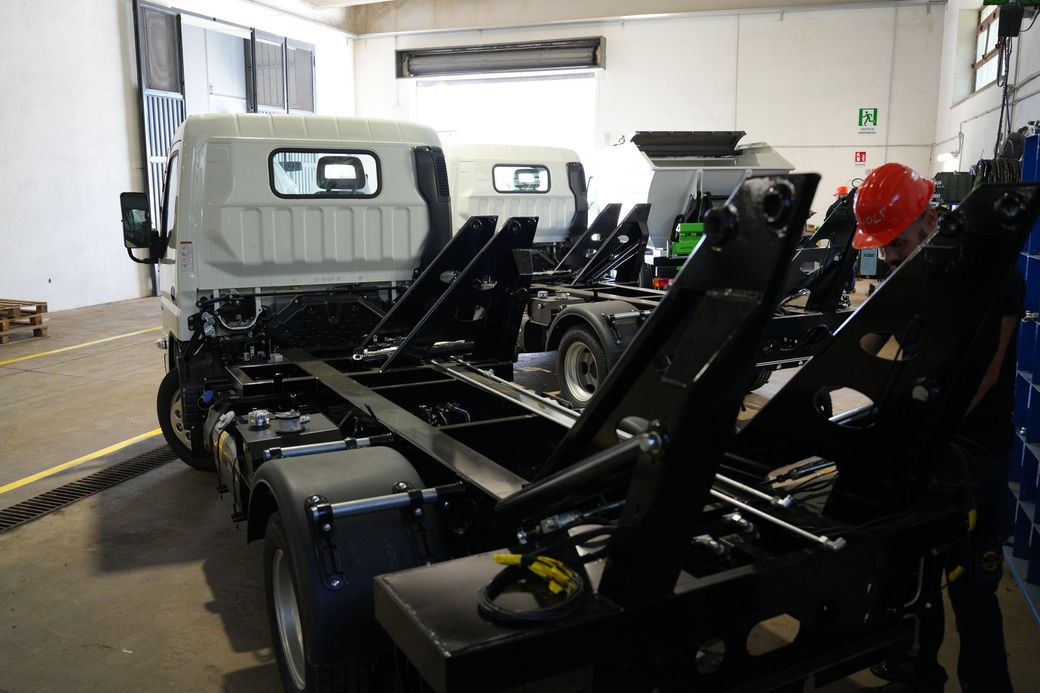 Fila di carrelli o macchinari industriali neri all'interno di un magazzino, con pannelli bianchi e una luce di sicurezza rossa.