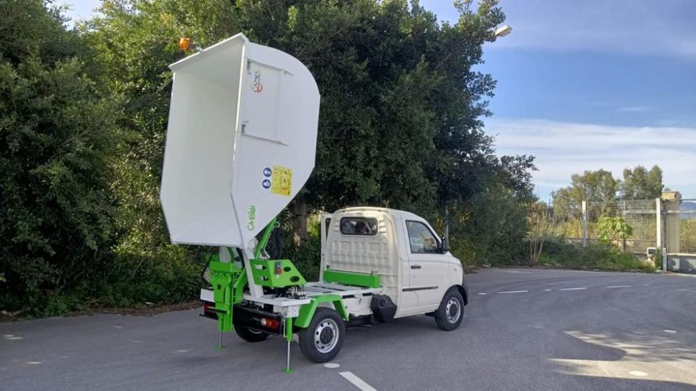 Un piccolo camioncino bianco con piattaforma elevatrice verde è parcheggiato su un piazzale asfaltato accanto ad alberi e cespugli.