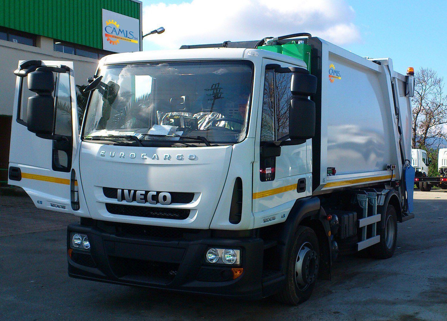 Un furgone Iveco bianco parcheggiato all'aperto vicino a un magazzino, rivolto verso la telecamera.
