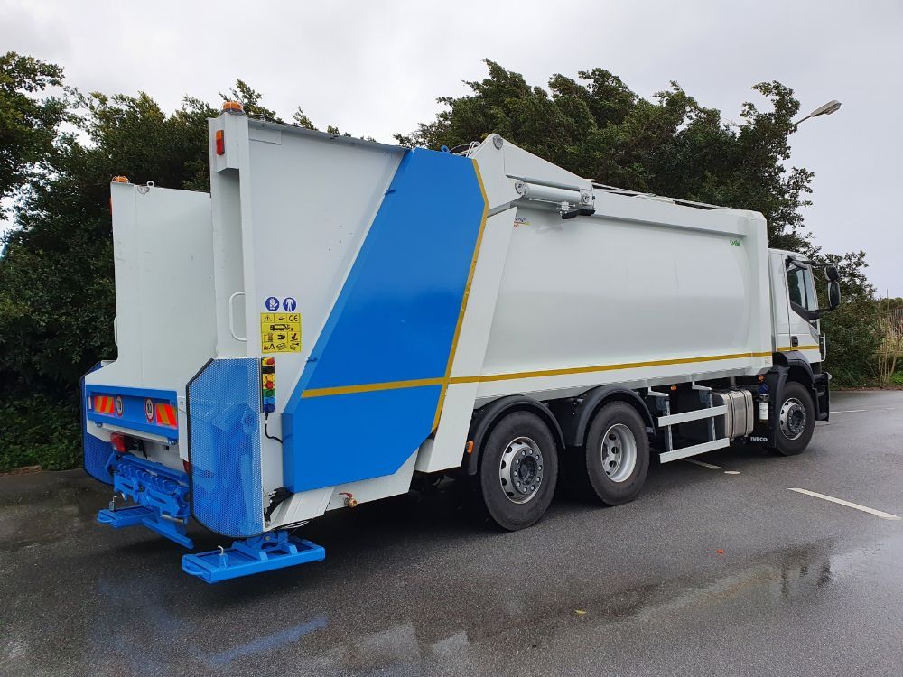 Camion della spazzatura bianco e blu parcheggiato su una strada bagnata accanto agli alberi.