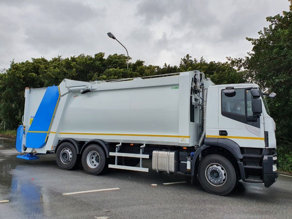 Un camion della spazzatura bianco su una strada bagnata, parcheggiato accanto alla vegetazione sotto un cielo nuvoloso.