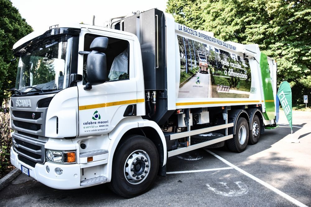 Un camion della spazzatura bianco parcheggiato in strada, con un pannello pubblicitario laterale e alberi sullo sfondo.
