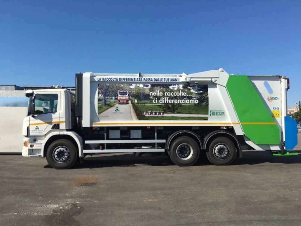 Un camion della spazzatura bianco con dettagli verdi parcheggiato sull'asfalto sotto un cielo azzurro.