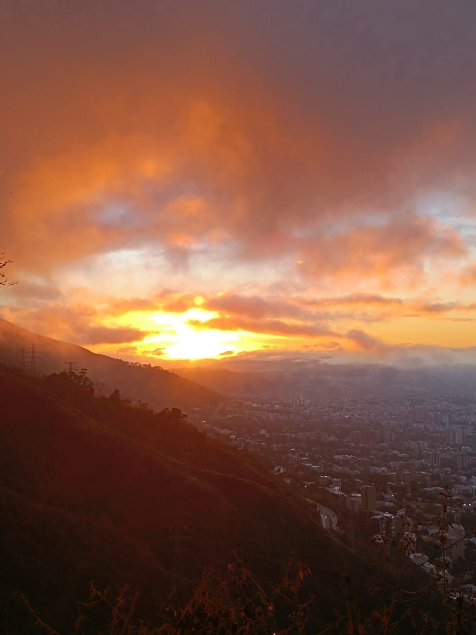 Sunset over a city, viewed from a mountain. Orange and yellow light illuminates clouds.