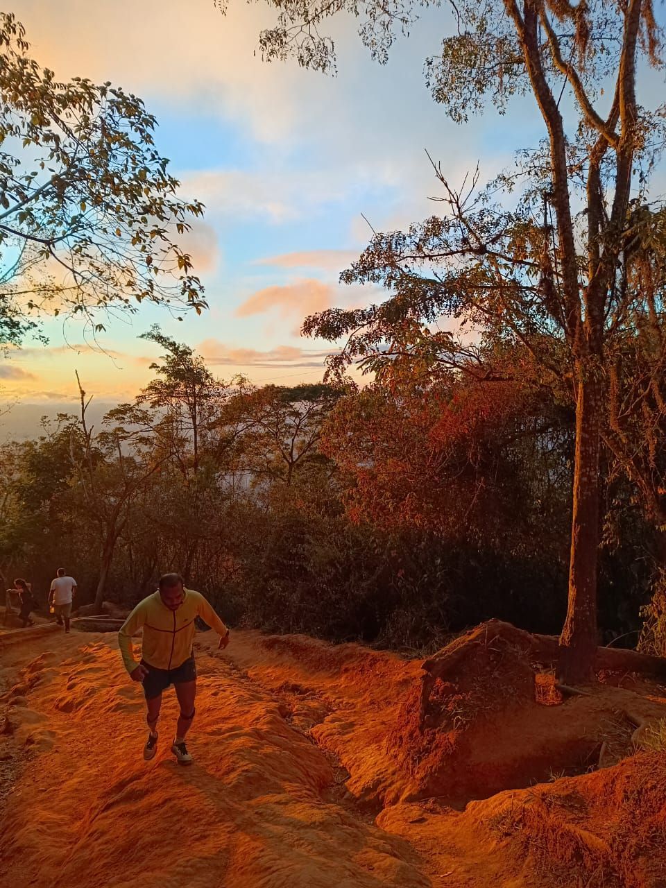 Man hiking on a dirt path at sunset. Orange light bathes the landscape, with trees and a colorful sky in the background.