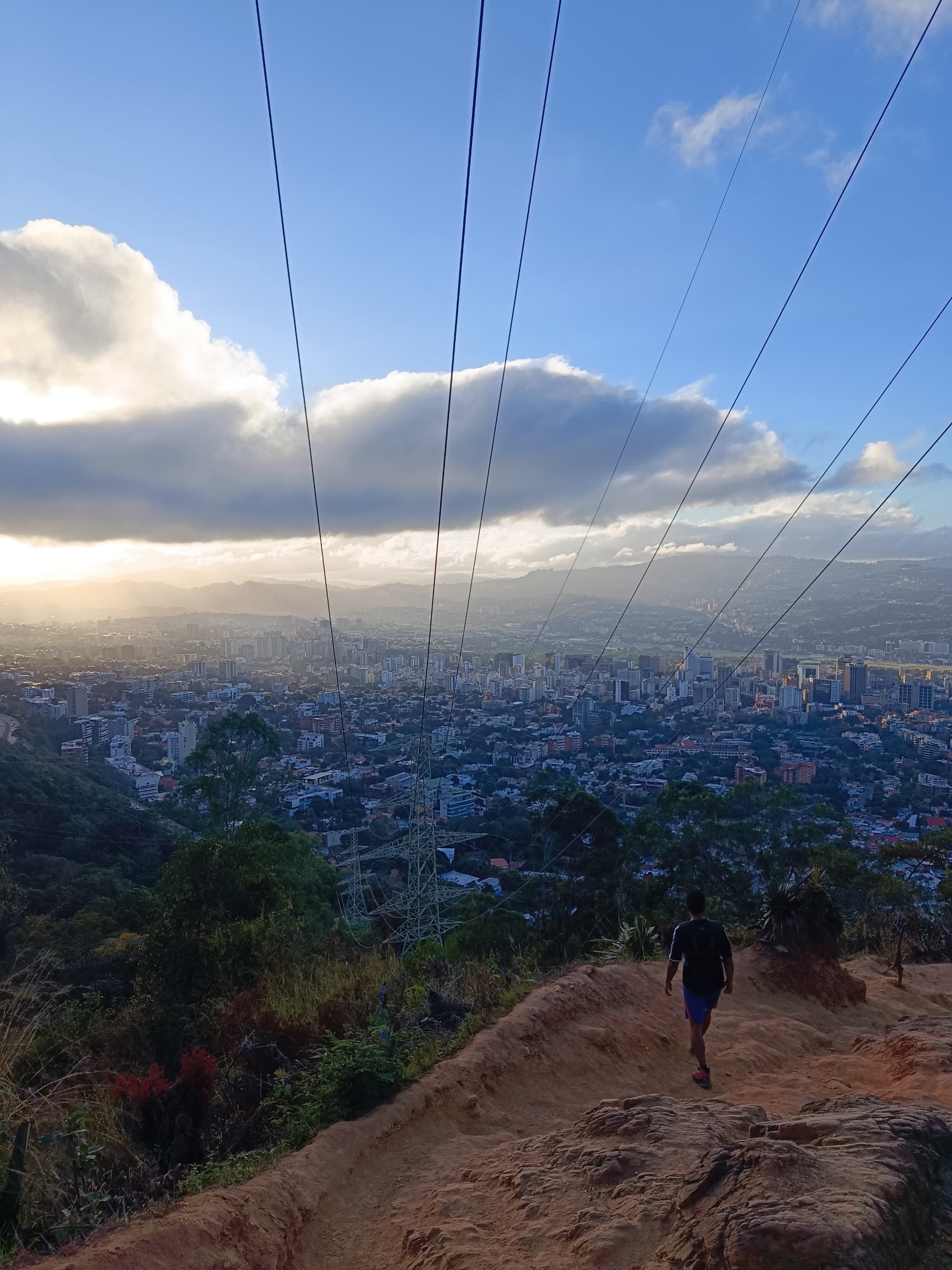 Person hikes on a hilltop overlooking a cityscape under a cloudy sky. Sunlight streams through clouds.