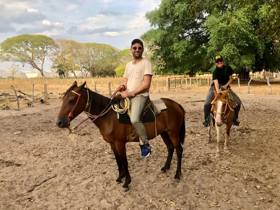 Two men on horseback in an outdoor setting. The man in front wears a light shirt and jeans; the other man is behind him.
